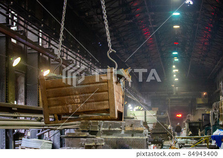 Big rusty metal container hanging from chains, lifted by a crane inside the foundry. Close-up with selective focus and background blur. Big rusty metal container hanging from chains, lifted by a crane inside the foundry. Close-up with selective focus and background blur. 84943400