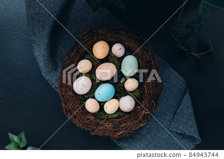 Top view wicker nest with moss and colored Easter eggs on dark background with green leaves. Happy Easter holiday. Muted colors. Flat lay, selective focus. Copy space 84943744