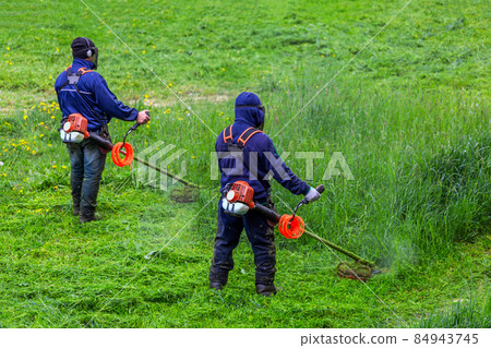 two lawnmower men with string trimmer and face mask trimmong grass - close-up 84943745