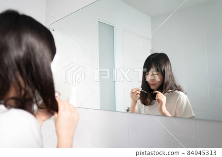 Young beautiful asian woman applying nutritional oils hair serum on her long black hair for treatment damaged hair in front of the bathroom mirror. Hair care concept. Young beautiful asian woman applying nutritional oils hair serum on her long black hair for treatment damaged hair in front of the bathroom mirror. Hair care concept. 84945313