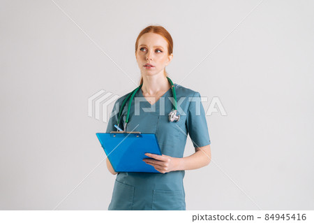 Portrait of thoughtful young female doctor in medical uniform writing prescription on clipboard, fills out medical history, looking away 84945416