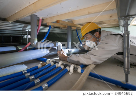 A man in work clothes inspecting the plumbing under the floor of a house A man in work clothes inspecting the plumbing under the floor of a house 84945692