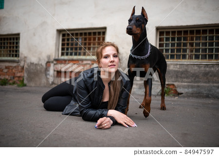 A girl in leather, with a Doberman, in an old yard. A girl in leather, with a Doberman, in an old yard. 84947597