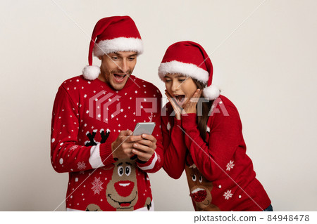 Surprised Couple In Cristmas Sweaters And Santa Hats Looking At Smartphone Screen 84948478