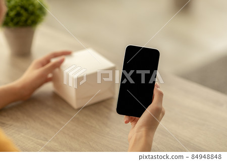 Woman holding cellphone with blank screen for mockup, writing review for product, touching cardboard box Woman holding cellphone with blank screen for mockup, writing review for product, touching cardboard box 84948848