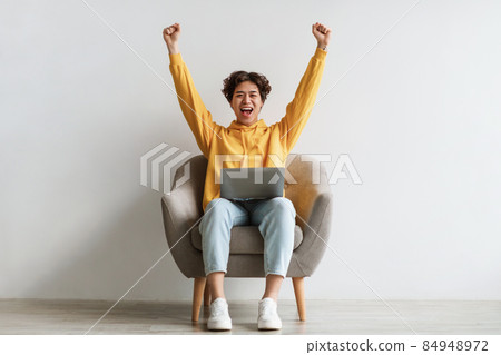 Young Asian guy sitting in armchair with laptop, raising hands up in excitement, celebrating success against white wall Young Asian guy sitting in armchair with laptop, raising hands up in excitement, celebrating success against white wall 84948972