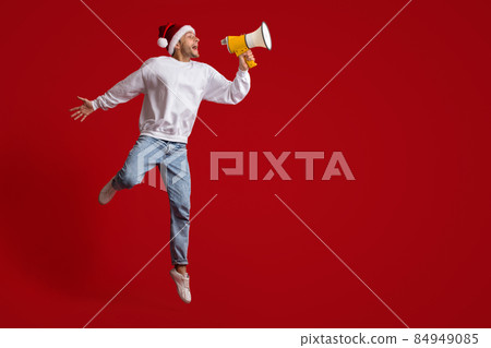 Christmas Announcement. Excited Young Guy In Santa Hat Shouting With Loudspeaker Christmas Announcement. Excited Young Guy In Santa Hat Shouting With Loudspeaker 84949085