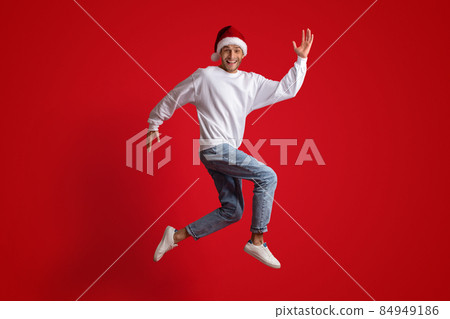 Holiday Fun. Cheerful Young Man Wearing Santa Hat Jumping Over Red Background 84949186