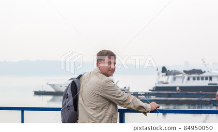 A man with a backpack in a wet jacket is waiting for his ship in the rain A man with a backpack in a wet jacket is waiting for his ship in the rain 84950050