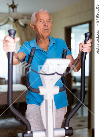 Old man using elliptic trainer at home Old man using elliptic trainer at home 84950109