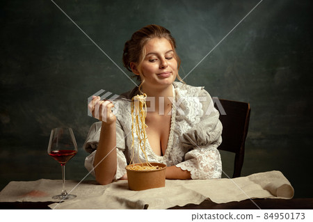 Half-length portrait of young beautiful girl in gray dress of medieval style having dinner isolated on dark vintage background. 84950173