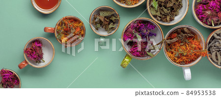 Assortment of dried relaxing tea herbs in colourful cups on mint green background. Calendula, mint, anise hyssop, monarda didyma, wormwood, sage leaves. 84953538