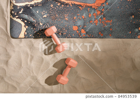 Unrolled grey yoga mat, orange dumbbells on sandy beach. Relaxation and meditation wellbeing. Mindful living concept 84953539
