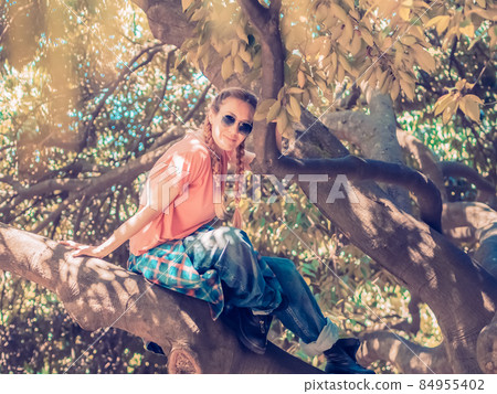 Smiling lady with two pigtails and a shirt tied on a belt sits on a tree in the park. Soft focus Smiling lady with two pigtails and a shirt tied on a belt sits on a tree in the park. Soft focus 84955402