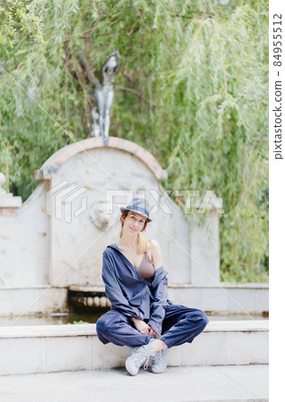 A casually dressed woman in a hat sits on the curb of a classic-style decorative park fountain A casually dressed woman in a hat sits on the curb of a classic-style decorative park fountain 84955512
