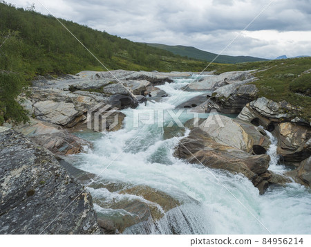Milky blue glacial Laddejahka river rapids with colorful stones in Lapland landscape with green mountains and birch trees at Padjelantaleden hiking trail, north Sweden wild nature. Summer cloudy day 84956214