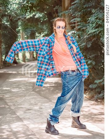 A girl in a plaid shirt with excessively long sleeves and huge jeans stands in a rough pose in a park on a green alley. A girl in a plaid shirt with excessively long sleeves and huge jeans stands in a rough pose in a park on a green alley. 84956227