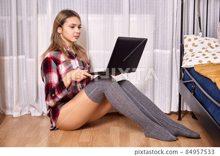 Thigh length socks on slender legs of young woman sitting on floor in bedroom with laptop on her lap. Thigh length socks on slender legs of young woman sitting on floor in bedroom with laptop on her lap. 84957533