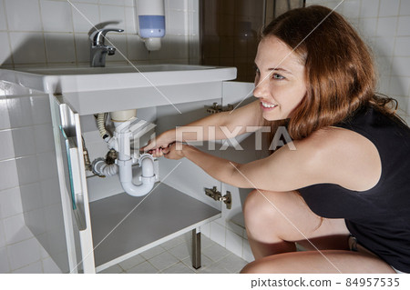 Woman tries to disassemble drain under sink to clear blockage. Woman tries to disassemble drain under sink to clear blockage. 84957535