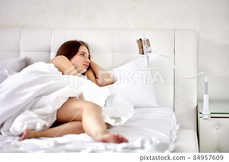 Smartphone holder with clamp is fixed on bedside table, woman is watching video of lie in bed. Smartphone holder with clamp is fixed on bedside table, woman is watching video of lie in bed. 84957609