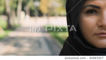 Half face portrait of muslim woman looking at camera, close-up. Arab woman pose in black hijab at city street. Serious, sad saudi female in traditional headscarf veil outdoors. With copy space text 84965957