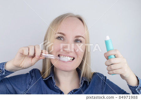 Close-up of a girl brushing teeth and look in front of a mirror. Oral hygiene. In the hands of objects for cleaning the oral cavity: a toothbrush and paste. Isolated on white background. Close-up of a girl brushing teeth and look in front of a mirror. Oral hygiene. In the hands of objects for cleaning the oral cavity: a toothbrush and paste. Isolated on white background. 84971988