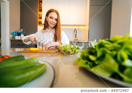 Portrait of young redhead woman cutting fresh yellow bell pepper preparing food salad sitting at table in modern kitchen room, looking at camera. Portrait of young redhead woman cutting fresh yellow bell pepper preparing food salad sitting at table in modern kitchen room, looking at camera. 84972997
