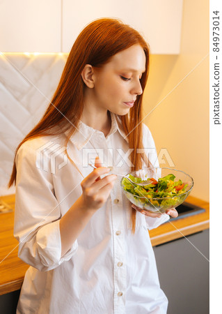 Vertical portrait of gorgeous attractive young redhead woman eating fresh vegetarian salad enjoying fresh tasty vegetables standing in kitchen room. Vertical portrait of gorgeous attractive young redhead woman eating fresh vegetarian salad enjoying fresh tasty vegetables standing in kitchen room. 84973014