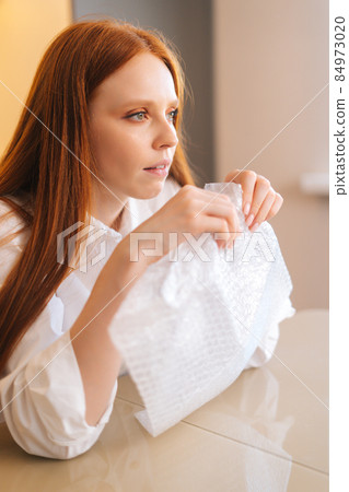 Vertical shot of serious young woman playing pops bubble wrap to calm herself sitting at table in kitchen. 84973020