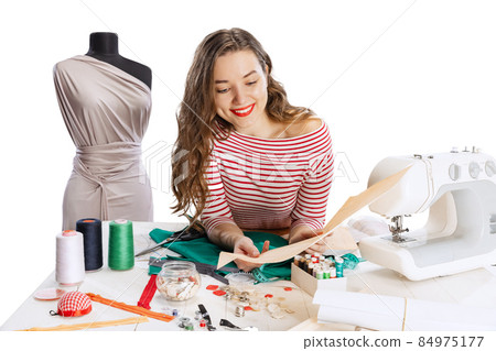 Cheerful beautiful young girl, seamstress, dressmaker working with fabric, scissors and sewing machine isolated on white background. Concept of job, hobby 84975177