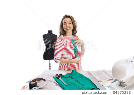 Cheerful beautiful young girl, seamstress, dressmaker working with fabric, scissors and sewing machine isolated on white background. Concept of job, hobby Cheerful beautiful young girl, seamstress, dressmaker working with fabric, scissors and sewing machine isolated on white background. Concept of job, hobby 84975228
