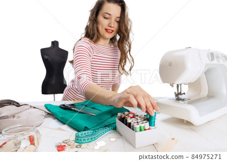 Cheerful beautiful young girl, seamstress, dressmaker working with fabric, scissors and sewing machine isolated on white background. Concept of job, hobby Cheerful beautiful young girl, seamstress, dressmaker working with fabric, scissors and sewing machine isolated on white background. Concept of job, hobby 84975271
