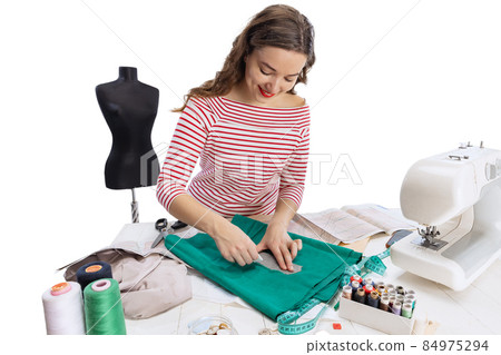 Cheerful beautiful young girl, seamstress, dressmaker working with fabric, scissors and sewing machine isolated on white background. Concept of job, hobby Cheerful beautiful young girl, seamstress, dressmaker working with fabric, scissors and sewing machine isolated on white background. Concept of job, hobby 84975294