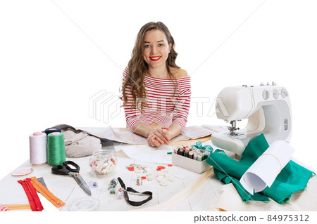 Cheerful beautiful young girl, seamstress, dressmaker working with fabric, scissors and sewing machine isolated on white background. Concept of job, hobby 84975312