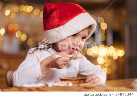 Little girl in red santa hat eating honey using honey dipper Kitchen decorated by fairy lights. Christmas mood at home. Happy New Year Little girl in red santa hat eating honey using honey dipper Kitchen decorated by fairy lights. Christmas mood at home. Happy New Year 84975344