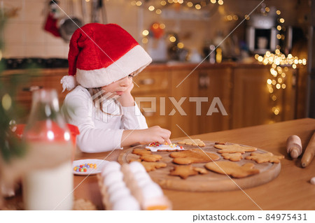 Little girl in santa hat decorates gingerbread using multicolored beads. Christmas and New Year traditions concept. Christmas bakery. Happy hollidays Little girl in santa hat decorates gingerbread using multicolored beads. Christmas and New Year traditions concept. Christmas bakery. Happy hollidays 84975431