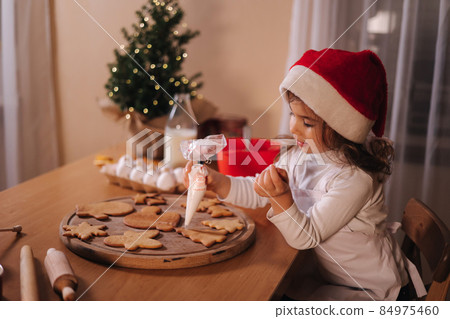 Little girl in santa hat decorates gingerbread at home. Christmas and New Year traditions concept. Christmas bakery. Happy hollidays 84975460