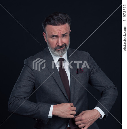Portrait of a stylish elegant senior businessman with a beard and casual business clothes in photo studio isolated on dark background gesturing with hands 84978755