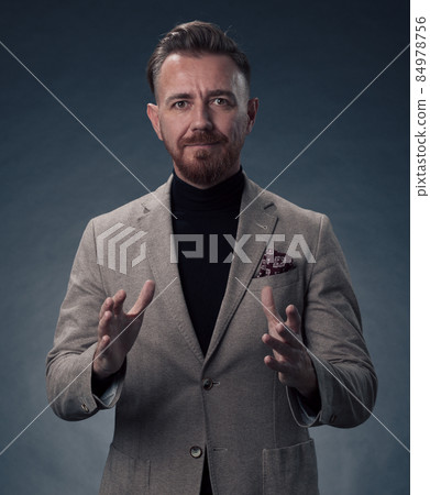 Portrait of a stylish elegant senior businessman with a beard and casual business clothes in photo studio isolated on dark background gesturing with hands 84978756