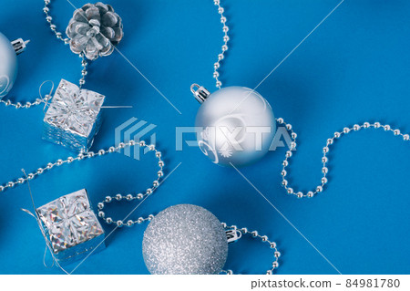 Silver baubles of Christmas decoration with golden lights on a blue background. Christmas balls, gift box, pine cones and new year's beads for the tree. Flat lay. 84981780