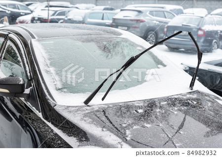 Close-up detail view of black modern car with raised up windshield wiper blades to prevent icing in cold frost winter day. Vehicle windscreen covered by whote snow parked on city street outdoors Close-up detail view of black modern car with raised up windshield wiper blades to prevent icing in cold frost winter day. Vehicle windscreen covered by whote snow parked on city street outdoors 84982932