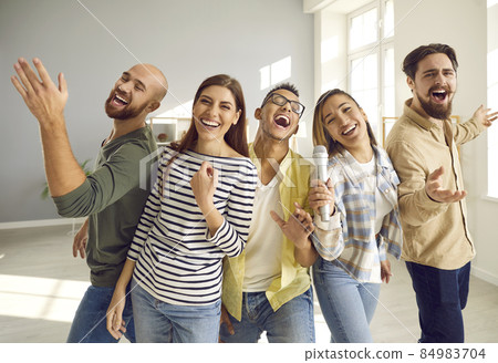 Young, cheerful and funny friends with microphone sing loudly and laugh while playing karaoke at home party. Close up of people in casual clothes. Concept of friendship, home entertainment and people. 84983704