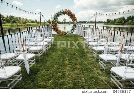 Round arch for a wedding ceremony by the river. White glass chairs stand for guests Round arch for a wedding ceremony by the river. White glass chairs stand for guests 84986337