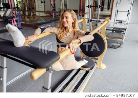 Cheerful young woman smiling while practicing pilates, having reformer training in stretching studio in the daytime Cheerful young woman smiling while practicing pilates, having reformer training in stretching studio in the daytime 84987107