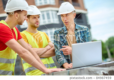 Engineer and two workers discussing near laptop outdoors Engineer and two workers discussing near laptop outdoors 84987459