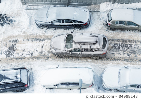 Top above aerial view of modern suv crossover car with roof rack box trunk driving through vehicles parked on city street covered by deep snow on clod frosty winter day. Apartment building driveway 84989148