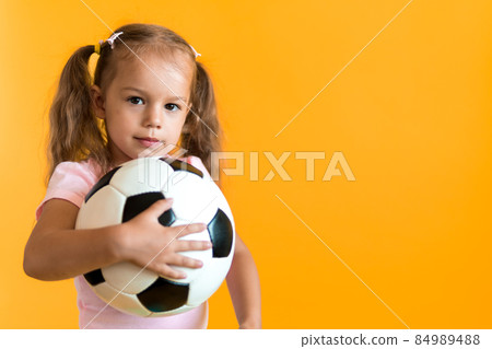 Authentic cute smiling preschool little girl with classic black and white soccer ball look at camera on yellow background. child play football in t-shirt and shorts. Sport, championship, team concept Authentic cute smiling preschool little girl with classic black and white soccer ball look at camera on yellow background. child play football in t-shirt and shorts. Sport, championship, team concept 84989488