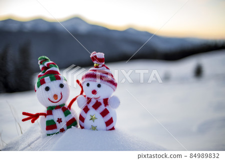 Two small funny toys baby snowman in knitted hats and scarves in deep snow outdoors on blurred mountains landscape background. Happy New Year and Merry Christmas greeting card theme. Two small funny toys baby snowman in knitted hats and scarves in deep snow outdoors on blurred mountains landscape background. Happy New Year and Merry Christmas greeting card theme. 84989832