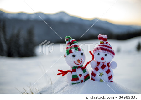 Two small funny toys baby snowman in knitted hats and scarves in deep snow outdoors on blurred mountains landscape background. Happy New Year and Merry Christmas greeting card theme. Two small funny toys baby snowman in knitted hats and scarves in deep snow outdoors on blurred mountains landscape background. Happy New Year and Merry Christmas greeting card theme. 84989833