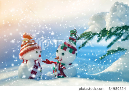 Two small funny toys baby snowman in knitted hats and scarves in deep snow outdoors near pine tree branch. Happy New Year and Merry Christmas greeting card. 84989834